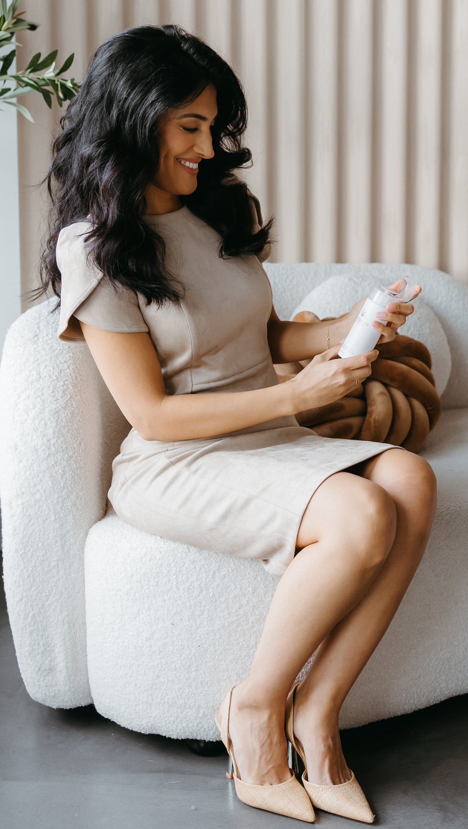 Business women with long dark hair smiling looking at makeup remover bottle on white couch in neutral setting  with natural light